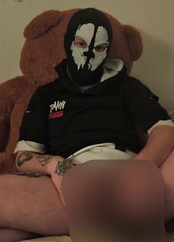 Download: Aloax - Eine kleine, maskierte Transperson spielt mit einem schwarzen Spielzeug und lehnt sich an ihren großen Teddybären.